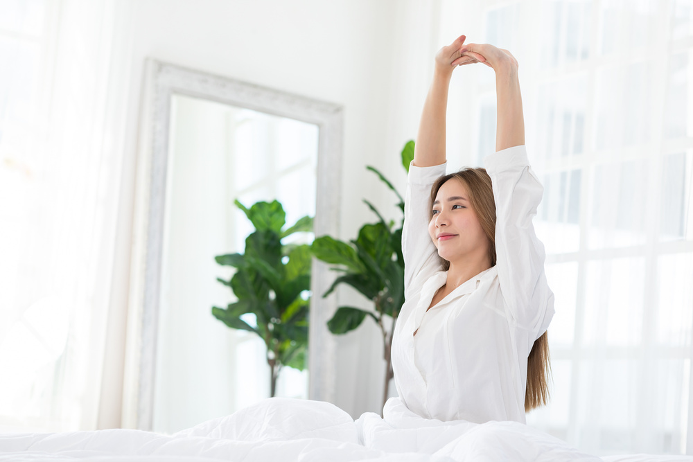 Beautiful Asian young Woman stretching her arm after wake up on bed and looking outside windows in cozy bedroom feeling so fresh and relax in the morning.Health care Concept
